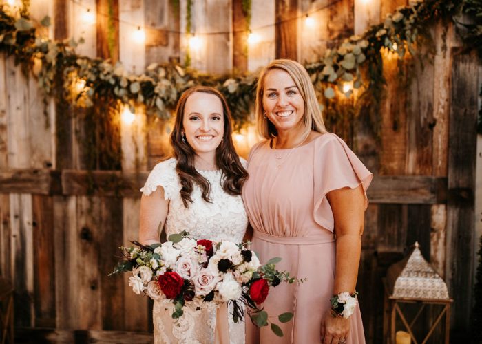 Mother of the Bride Standing with Bride Wearing Maggie Sottero Wedding Dress in Front of Rustic Wedding Backdrop with Hanging Lights