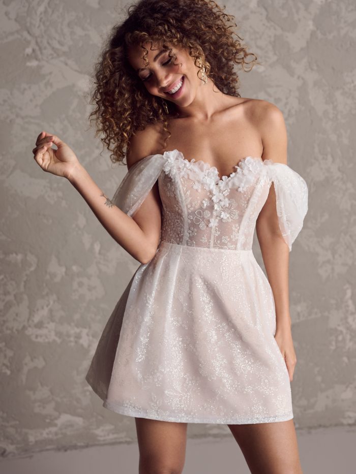 Bride wearing the Zoe short wedding dress featuring an off the shoulder floral appliqué bodice, sheer corset details, and a sparkly A-line mini skirt.