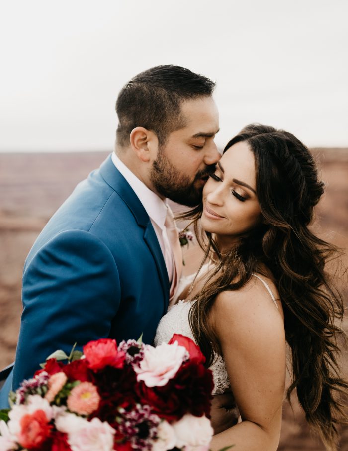 Groom in Moab Kissing Bride Wearing Rebecca Ingram Wedding Dress and Romantic Bridal Makeup