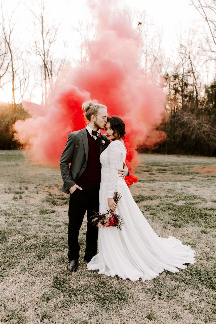 Groom Kissing Bride on Forehead with Red Powder Cannon Behind Them