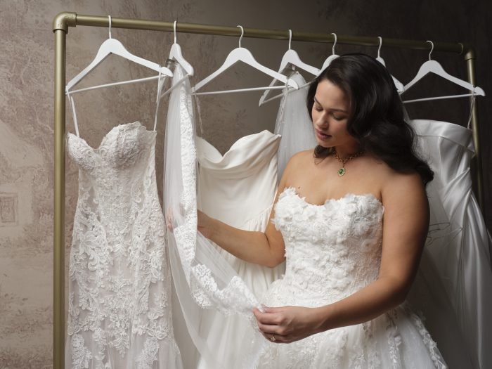 Bride browsing a rack of wedding dresses while wearing a strapless floral lace ballgown with 3D floral details.