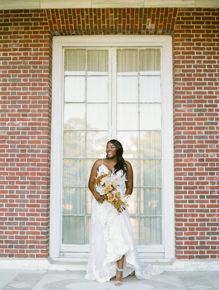 Bride Wearing Lace Boho Sheath Wedding Dress Called Tuscany Lane by Maggie Sottero in front of brick building