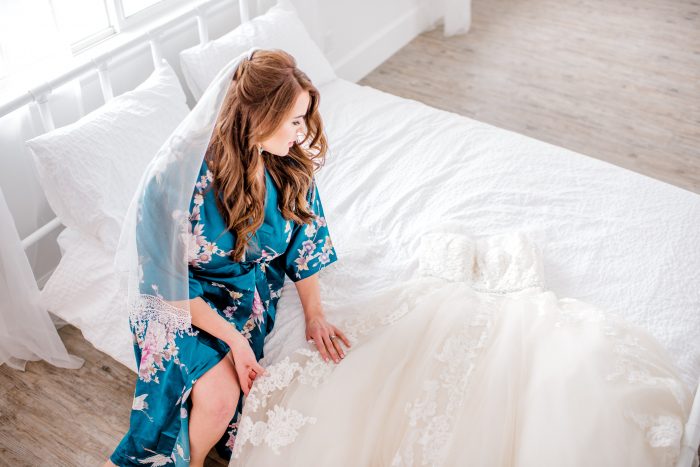 Bride in Robe on a Bed Looking at her Wedding Dress by Maggie Sottero Cover Photo