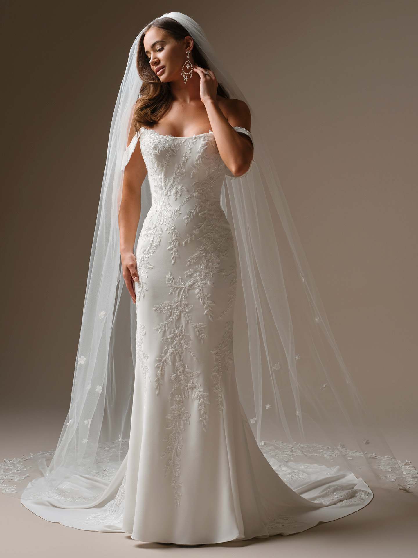 Thera sheath wedding dress featuring an off-the-shoulder neckline, fitted silhouette, and delicate floral lace appliqués with a soft, romantic train and sheer veil