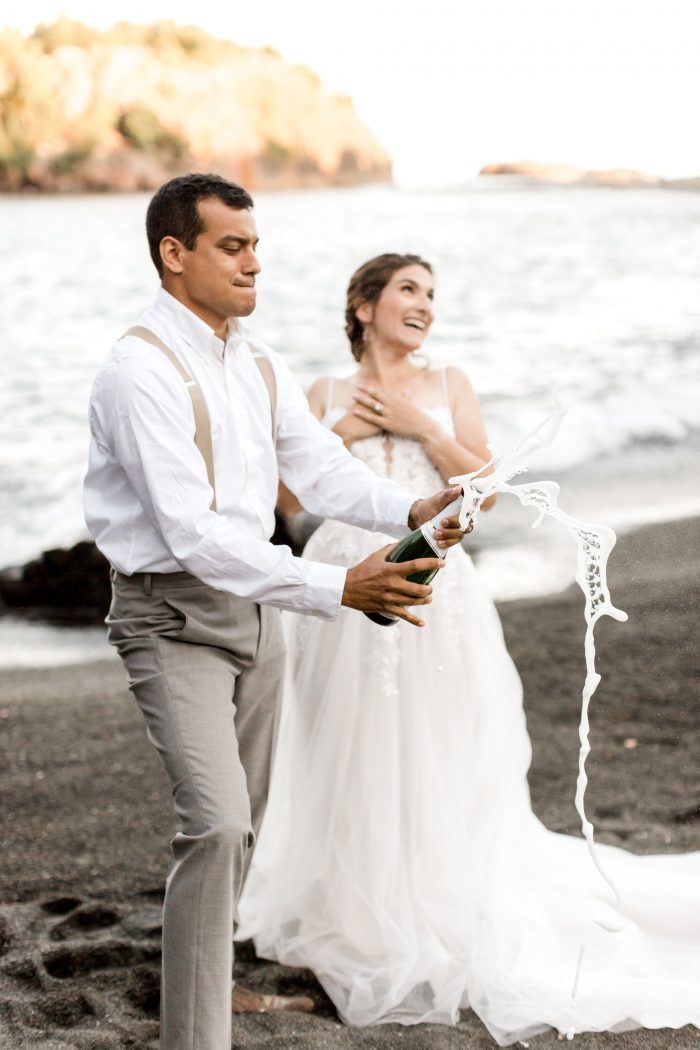 Groom Popping Open Bottle of Champagne on Beach for Wedding Toast While Bride Laughs