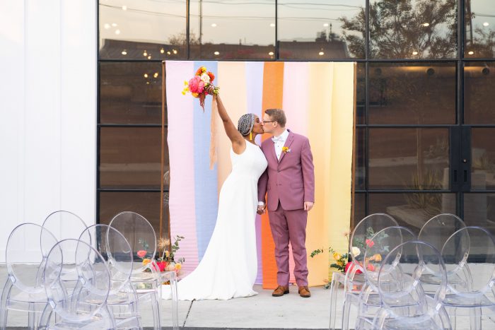 Groom Kissing Bride at Colorblock Wedding While Bride Raises Arm with Bouquet