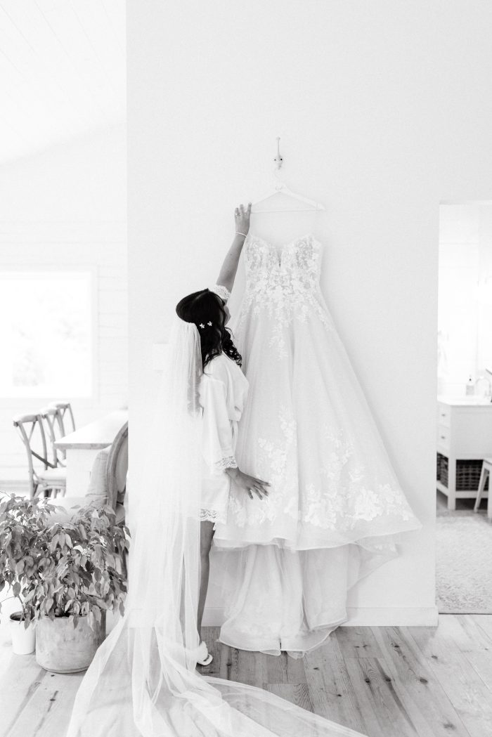 Bride In Bridal PJs Grabbing Casey Wedding Dress by Maggie Sottero off Hanger on the wall