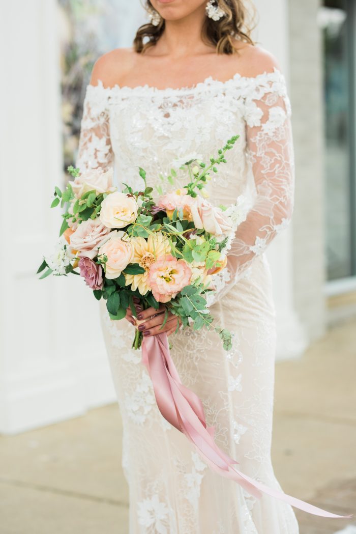Model holding pastel floral bouquet while wearing Emiliano sexy lace wedding dress by Maggie Sottero