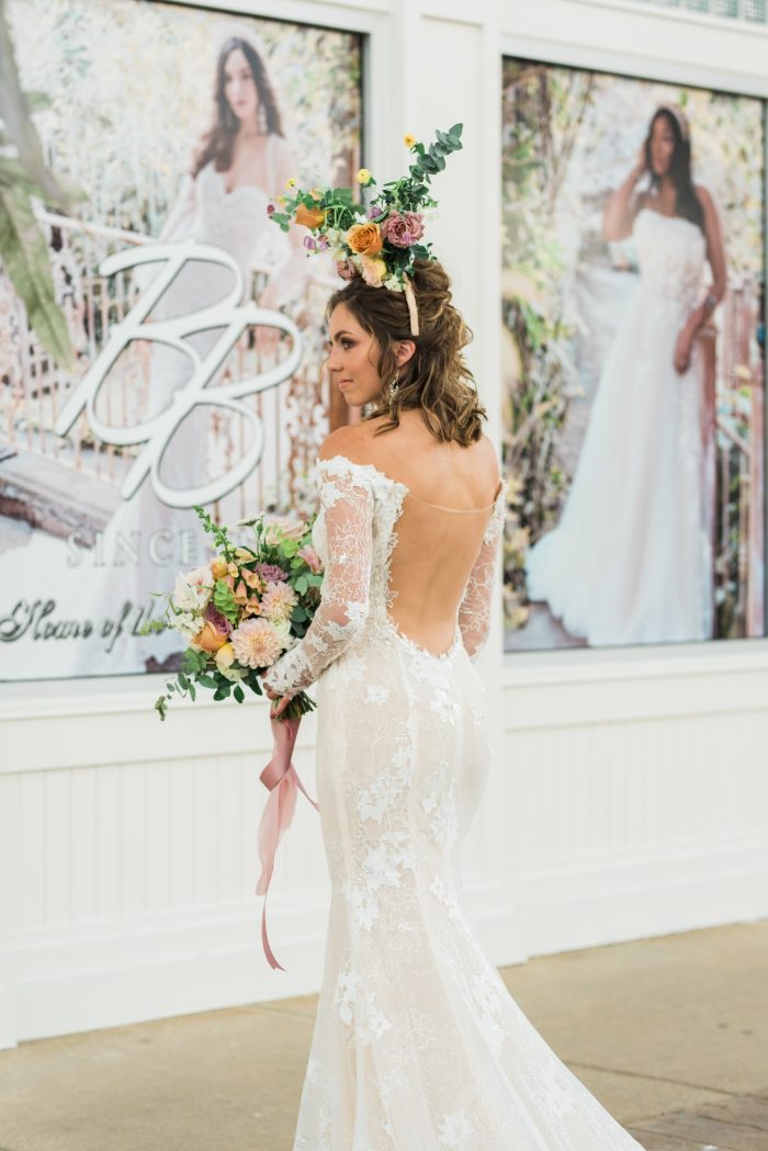 Model holding pastel floral bouquet while wearing Emiliano sexy backless lace wedding dress by Maggie Sottero