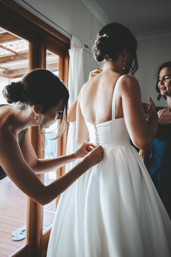 Bridal Party Helping Bride Get Ready by Buttoning Up Her Satin Wedding Dress Called Selena by Maggie Sottero