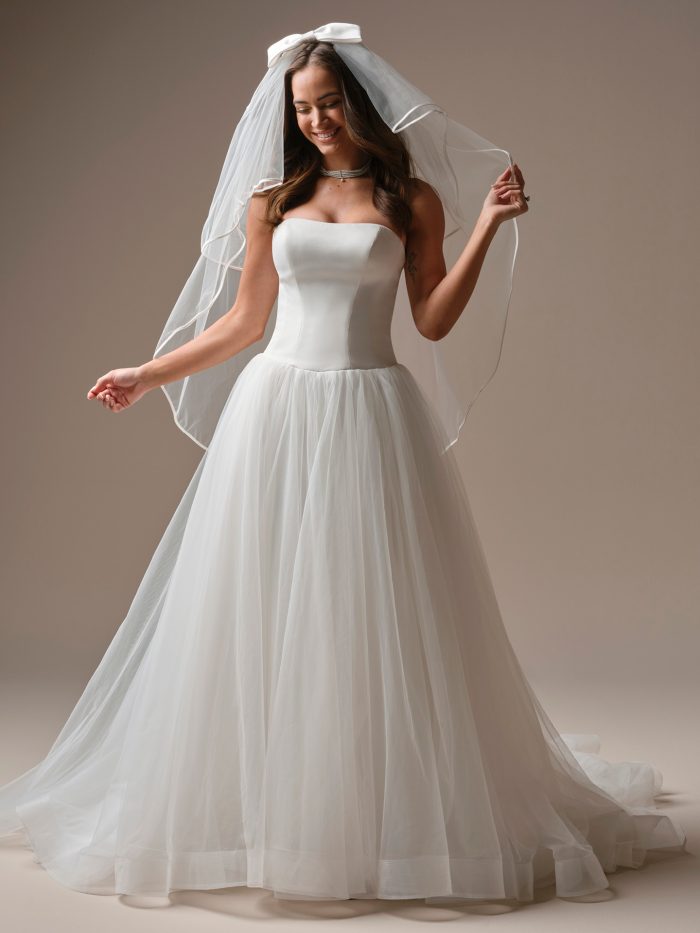 Bride wearing Maggie Sottero Hudson Fingertip Veil with ribbon edge paired with a strapless ballgown wedding dress, front view.