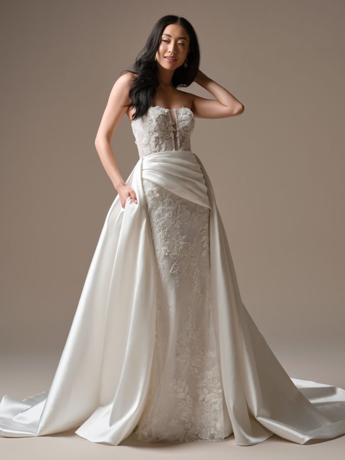 Bride wearing Maggie Sottero Laurie overskirt satin overskirt layered over a strapless lace wedding dress, full-length view with detachable overskirt.