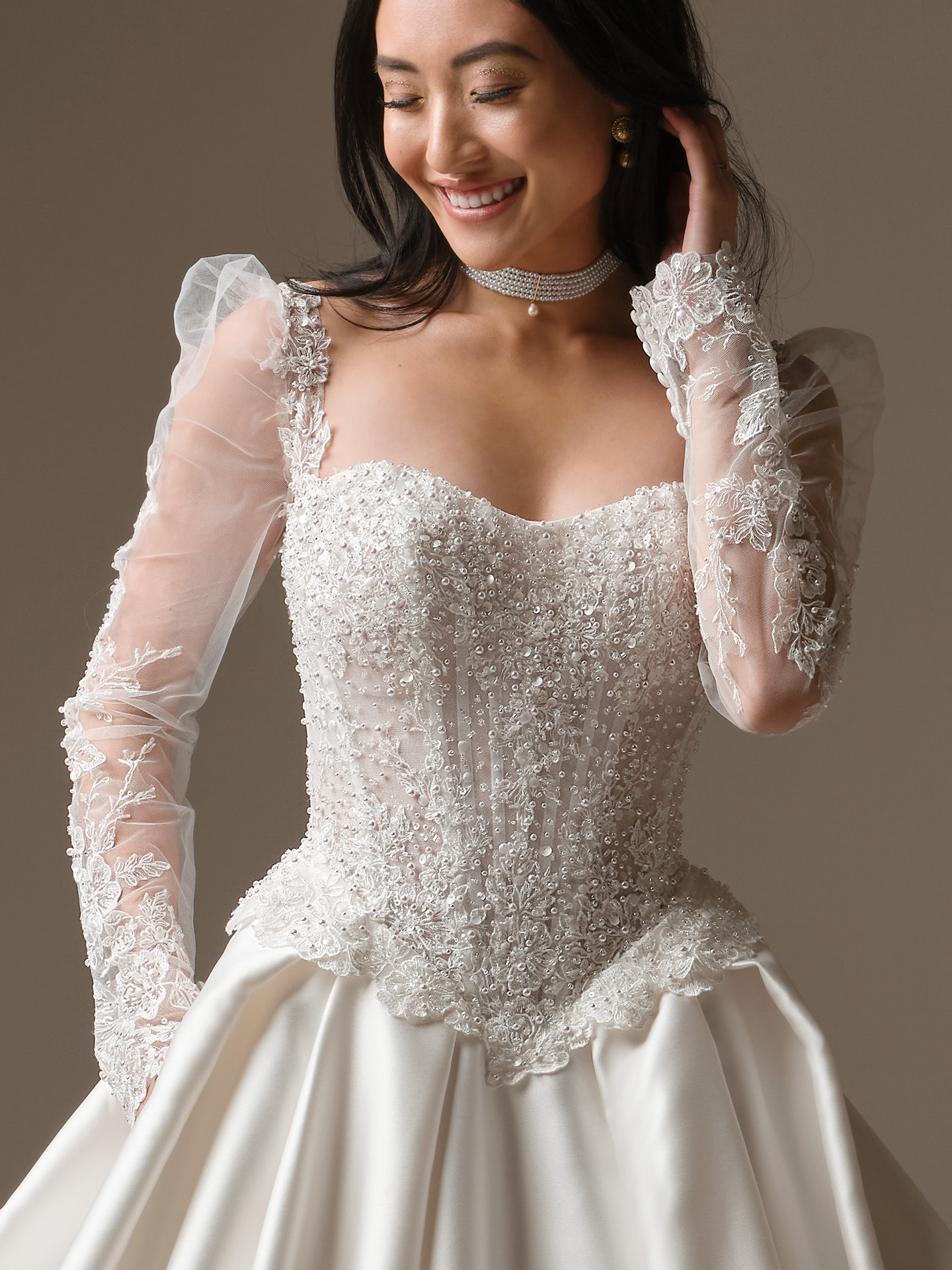 Bride wearing Maggie Sottero London sleeves with illusion lace long sleeves and beaded bodice paired with a satin ballgown wedding dress, close-up view.