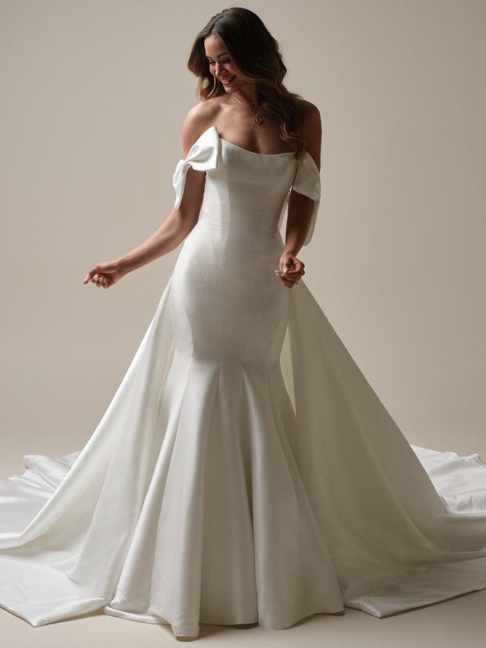 Bride wearing Sottero and Midgley Brennan fit-and-flare wedding dress with detachable cape, featuring a strapless satin gown with off-the-shoulder bow details and a dramatic flowing cape train, shown from front and back views.