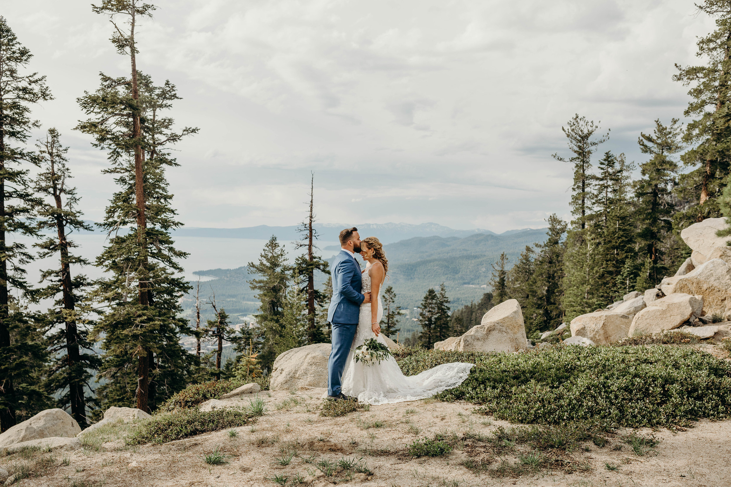 Mariée vêtue d'une robe appelée Toscane Lynette par Maggie Sottero debout au sommet des montagnes avec des arbres avec son mari