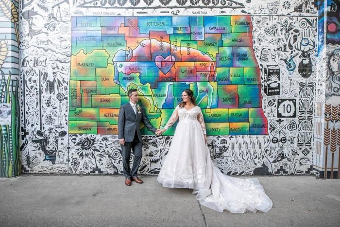 Bride Standing Next To Husband In Front Of Colorful Mural Wearing A Wedding Dress Called Zander By Sottero And Midgley