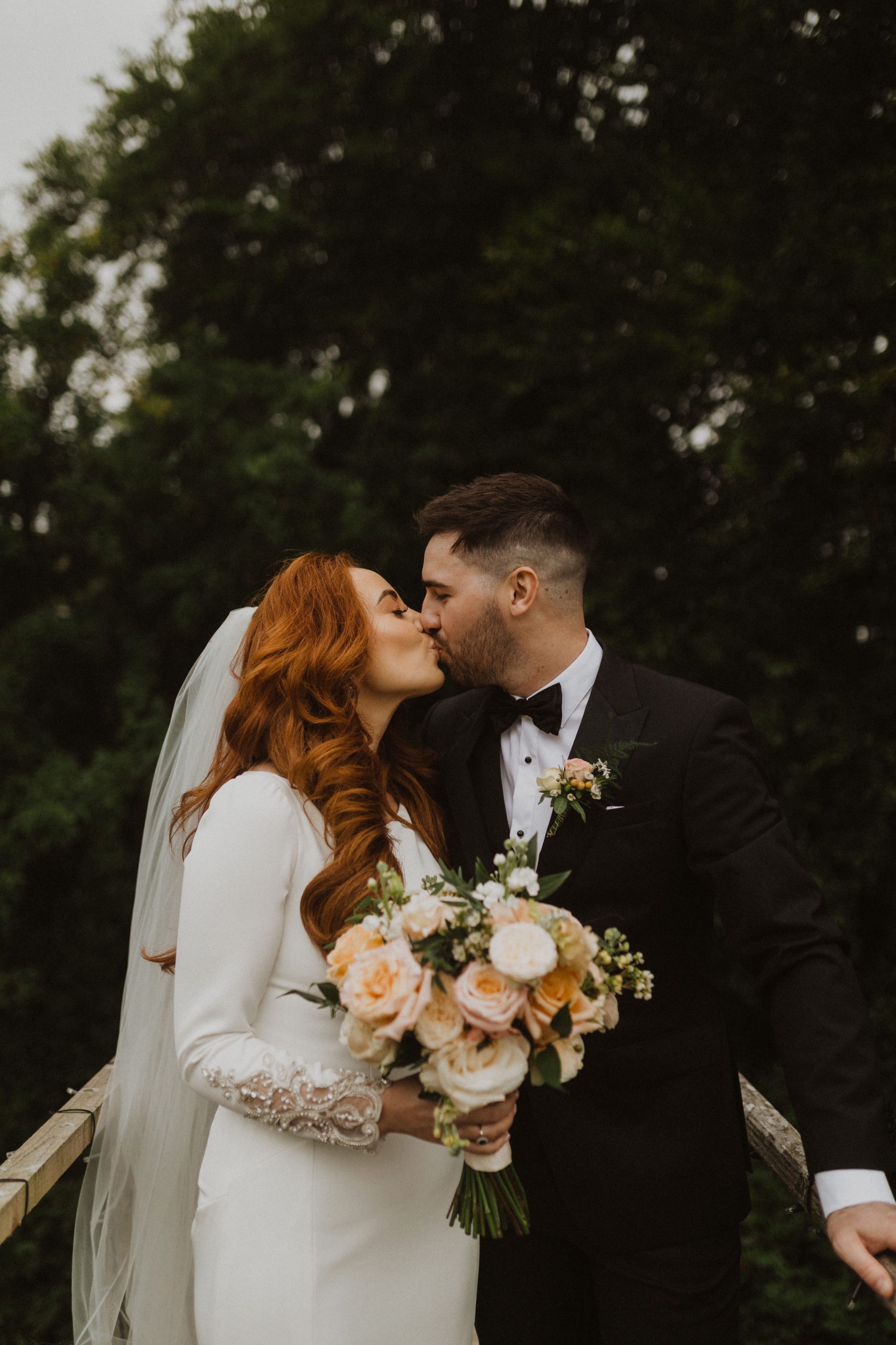 Mariée aux cheveux rouges embrassant son mari tout en tenant un bouquet aux couleurs de mariage de printemps vêtue d'une robe de mariée appelée Aston par Sottero et Midgley