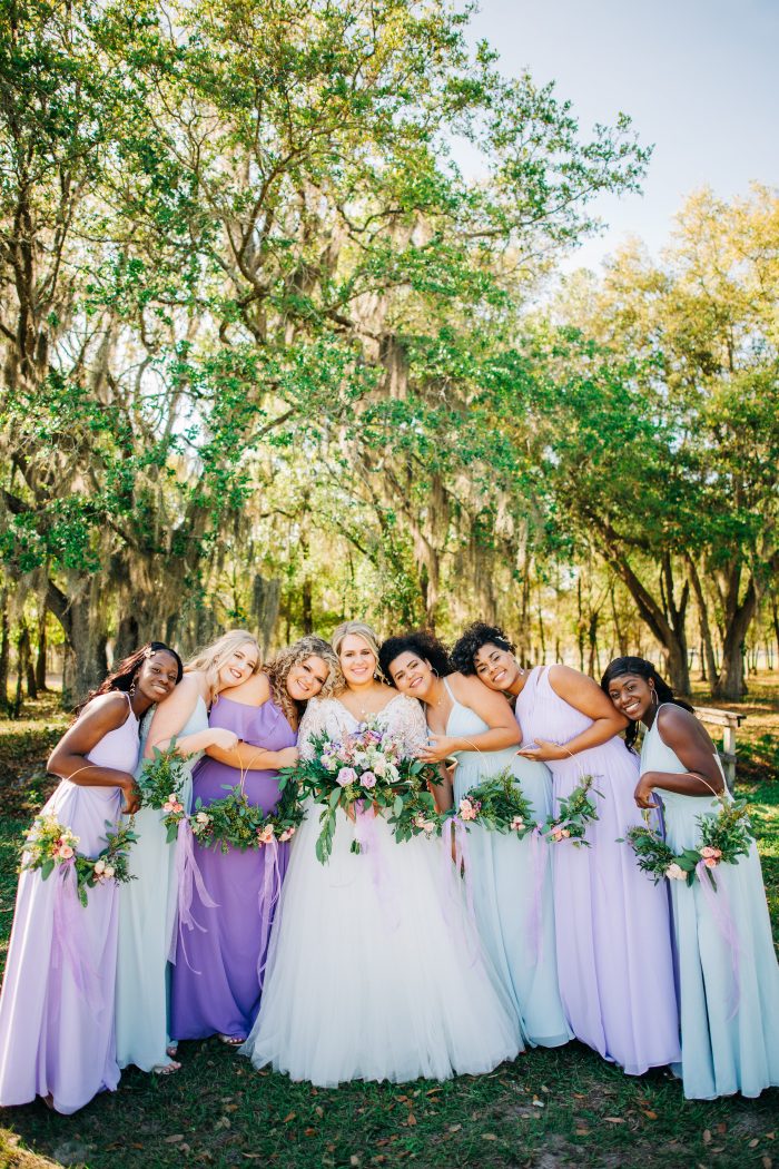 Bridal Party Wearing Spring Wedding Colors Of Purple And Blue Standing With Bride Wearing A Long Sleeved Wedding Gown Called Mallory Dawn By Maggie Sottero