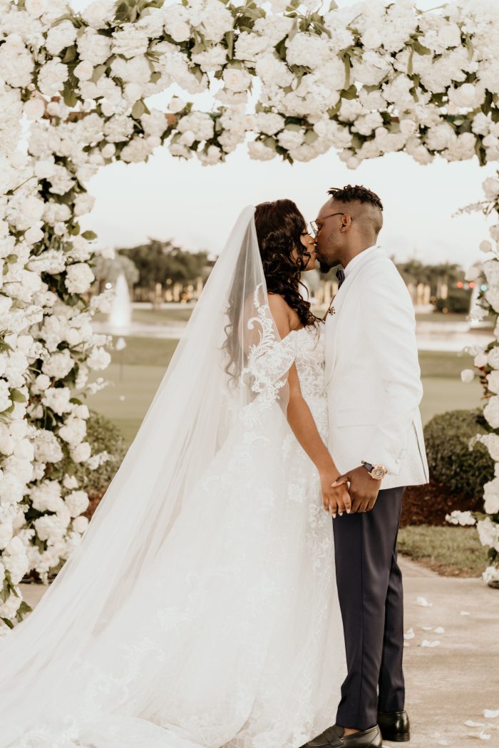Curvy Black Bride Wearing An A-Line Wedding Dress Called Parker By Maggie Sottero Standing Under Flower Arch With Black Groom