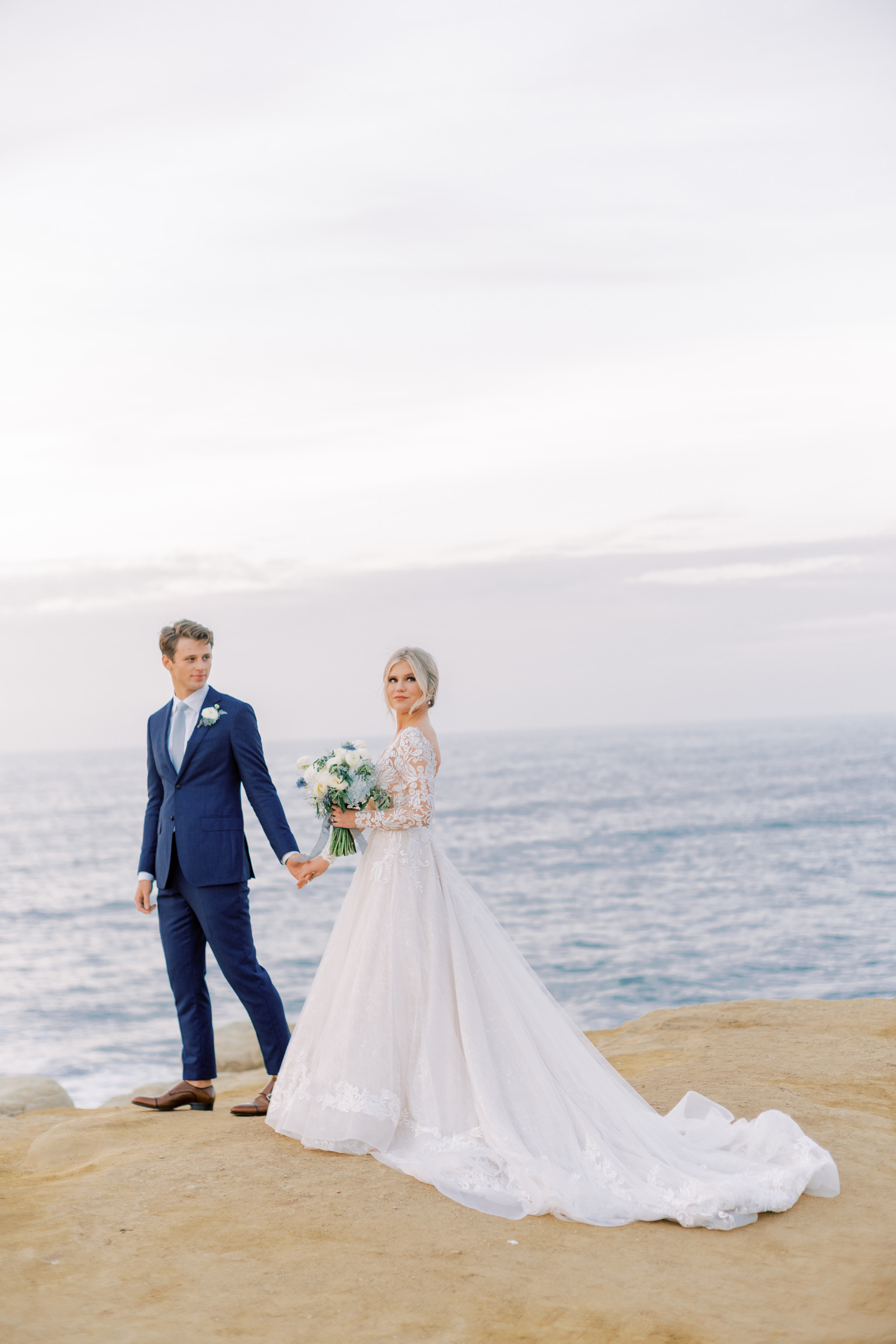 Mariée portant une robe de mariée en dentelle A-ligne avec des manches appelée Zander par Sottero et Midgley sur la falaise du bord de mer avec le marié