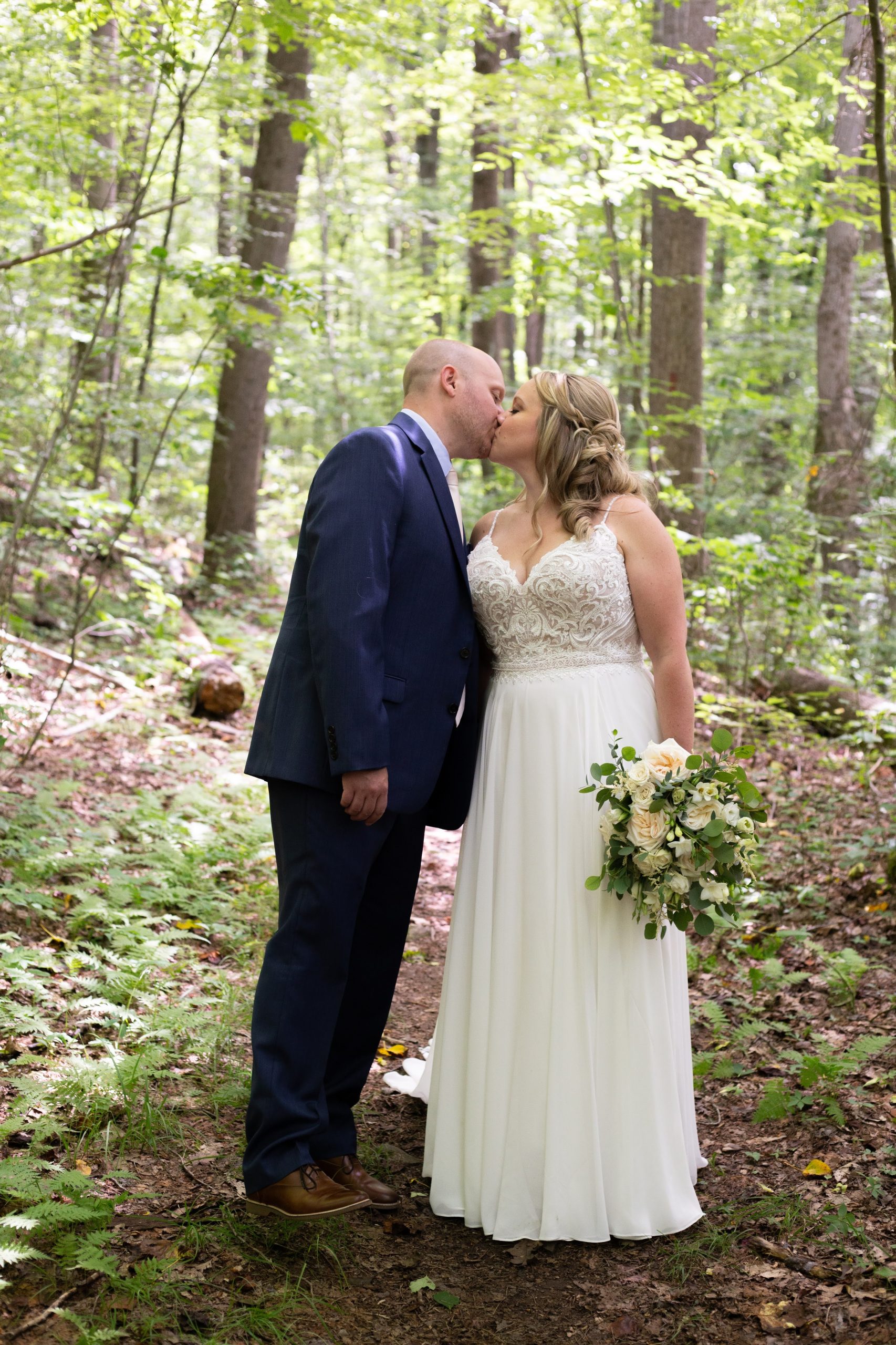 Mariée portant une robe de mariée de printemps appelée Lorraine par Maggie Sottero debout dans les bois avec le marié en robe de mousseline