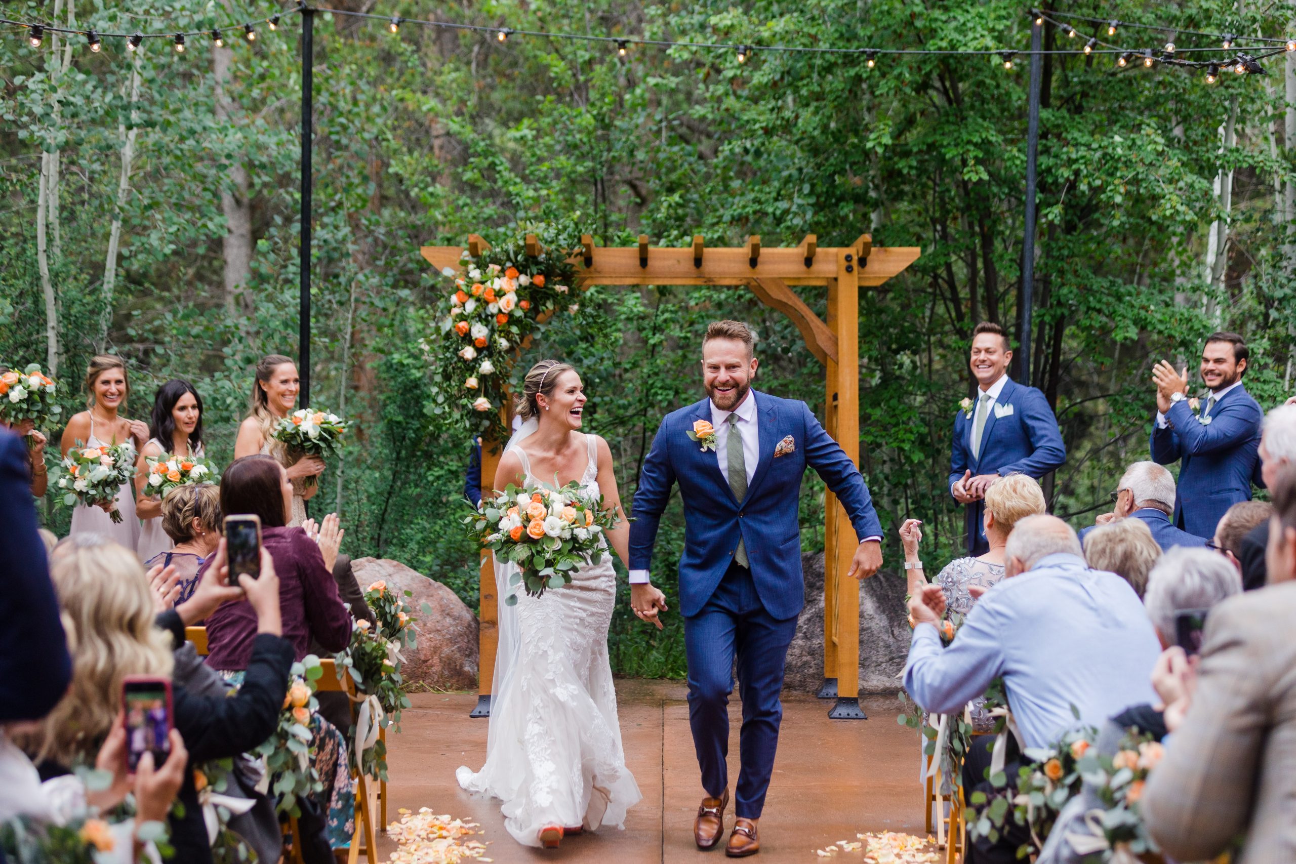 Bride Wearing Greenley By Maggie Sottero Walking Down The Aisle With Groom In Navy Blue Suit In Outdoor Forest Venue