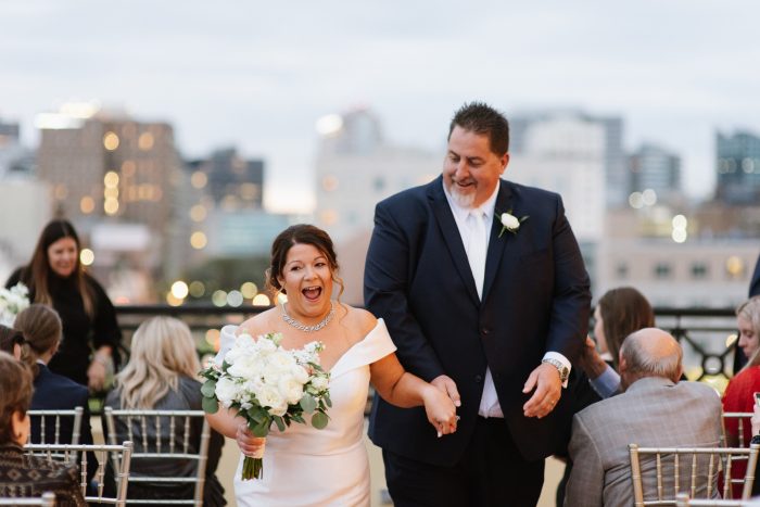 Bride Wearing An Off-The-Shoulder Wedding Dress Called Josie By Rebecca Ingram Walking Down The Aisle With Groom In Rooftop Ceremony