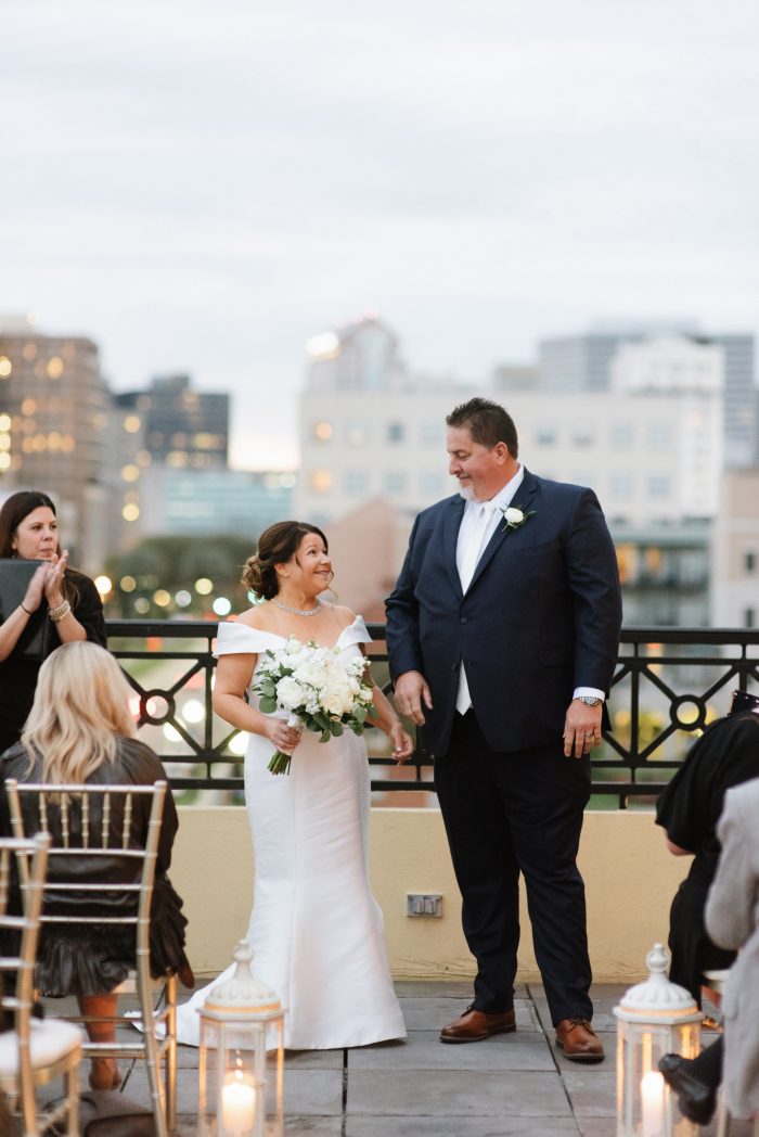 Bride Wearing An Off-The-Shoulder Wedding Dress Called Josie By Rebecca Ingram Walking Down The Aisle With Groom In Rooftop Ceremony