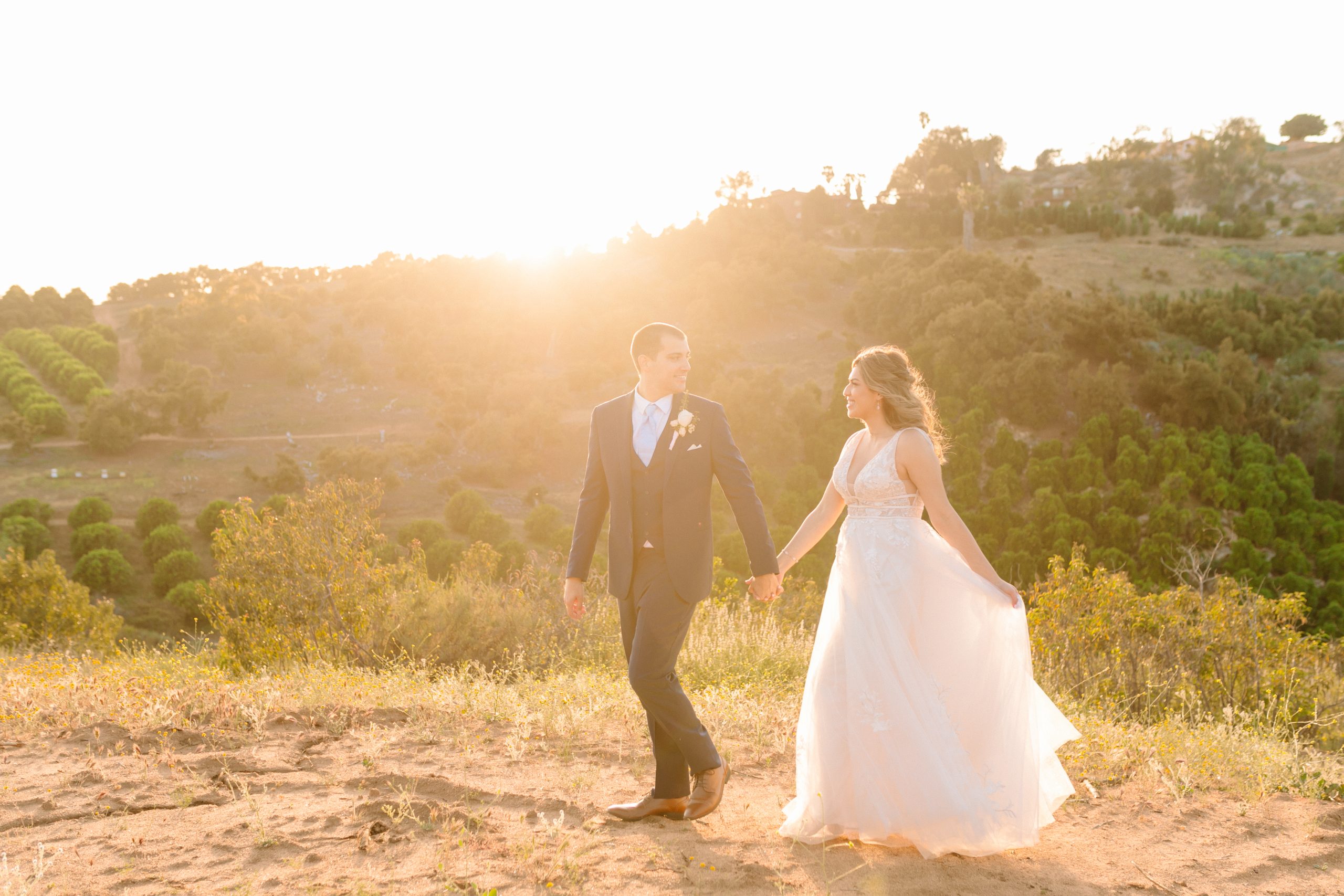 Photo de la mariée et du marié marchant au coucher du soleil avec la mariée portant une robe de mariée A-Line appelée Raelynn par Rebecca Ingram
