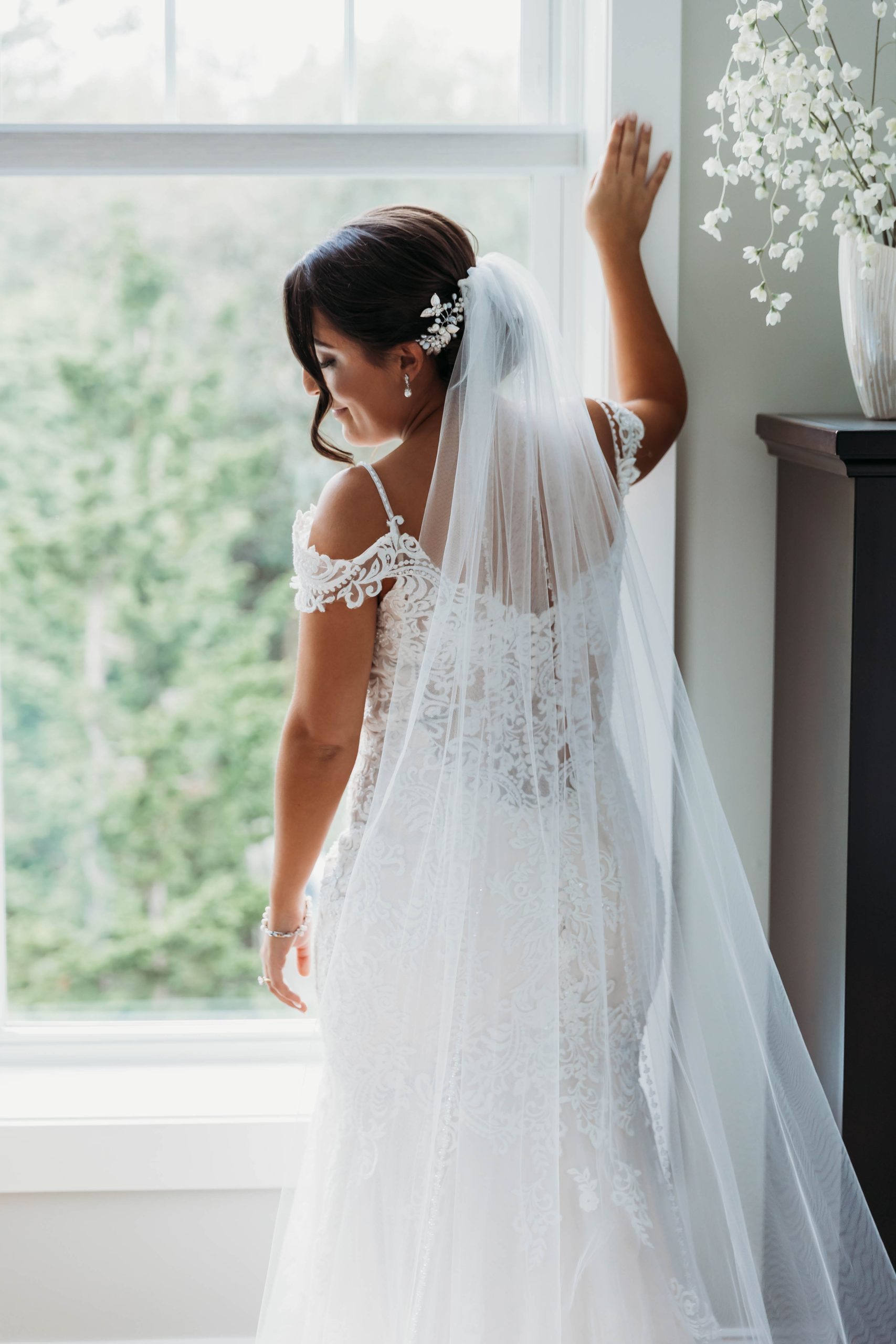 Mariée portant une robe de mariée en dentelle appelée Katell par Maggie Sottero devant la fenêtre