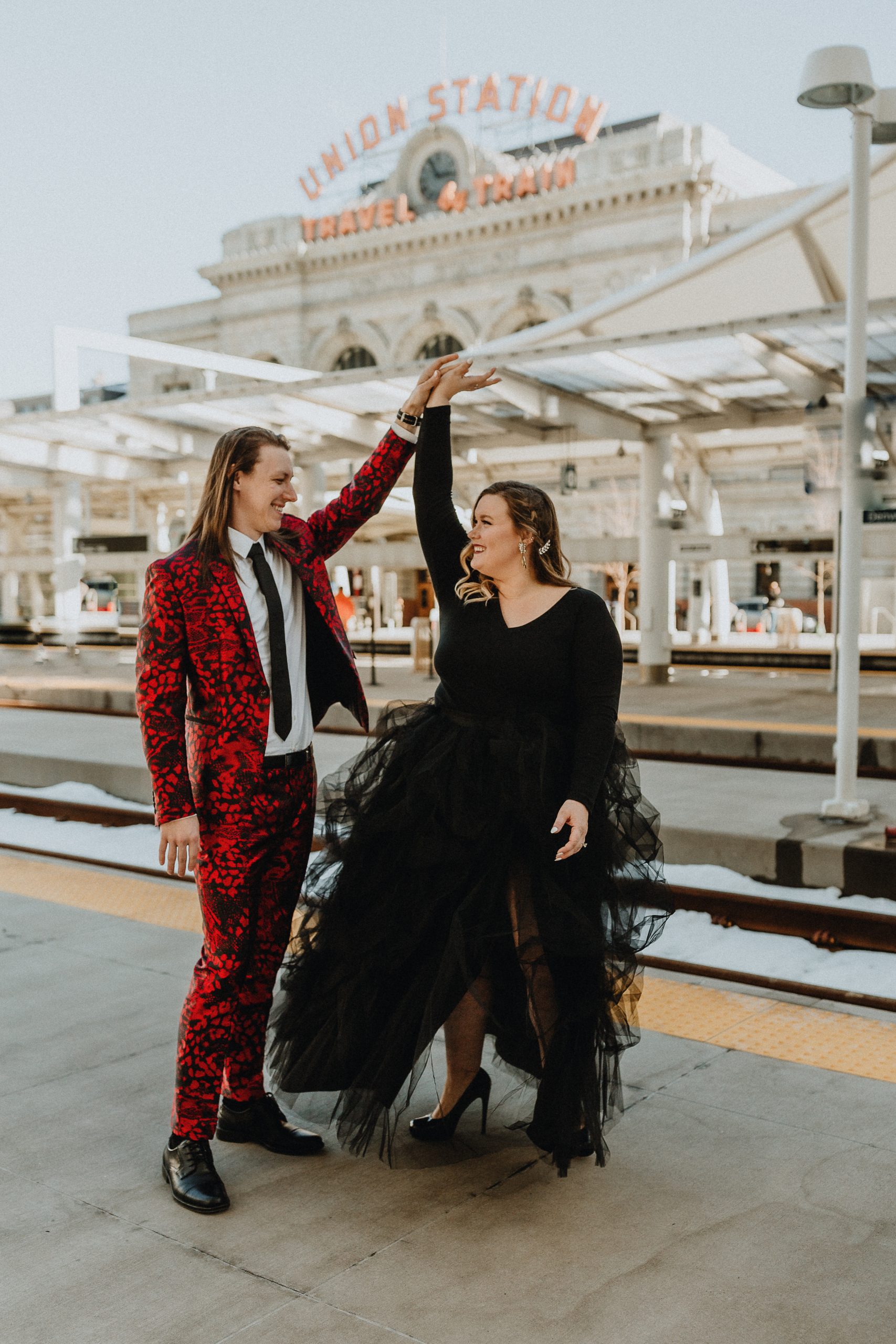 Mariée en robe noire avec un fiancé en costume rouge lors d'une séance photo de fiançailles à la gare Union de Denver