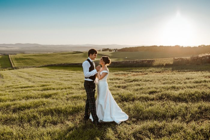 Bride And Groom Standing In A Field Wearing A Simple Off The Shoulder Wedding Dress Called Josie By Rebecca Ingram With Groom In A Field