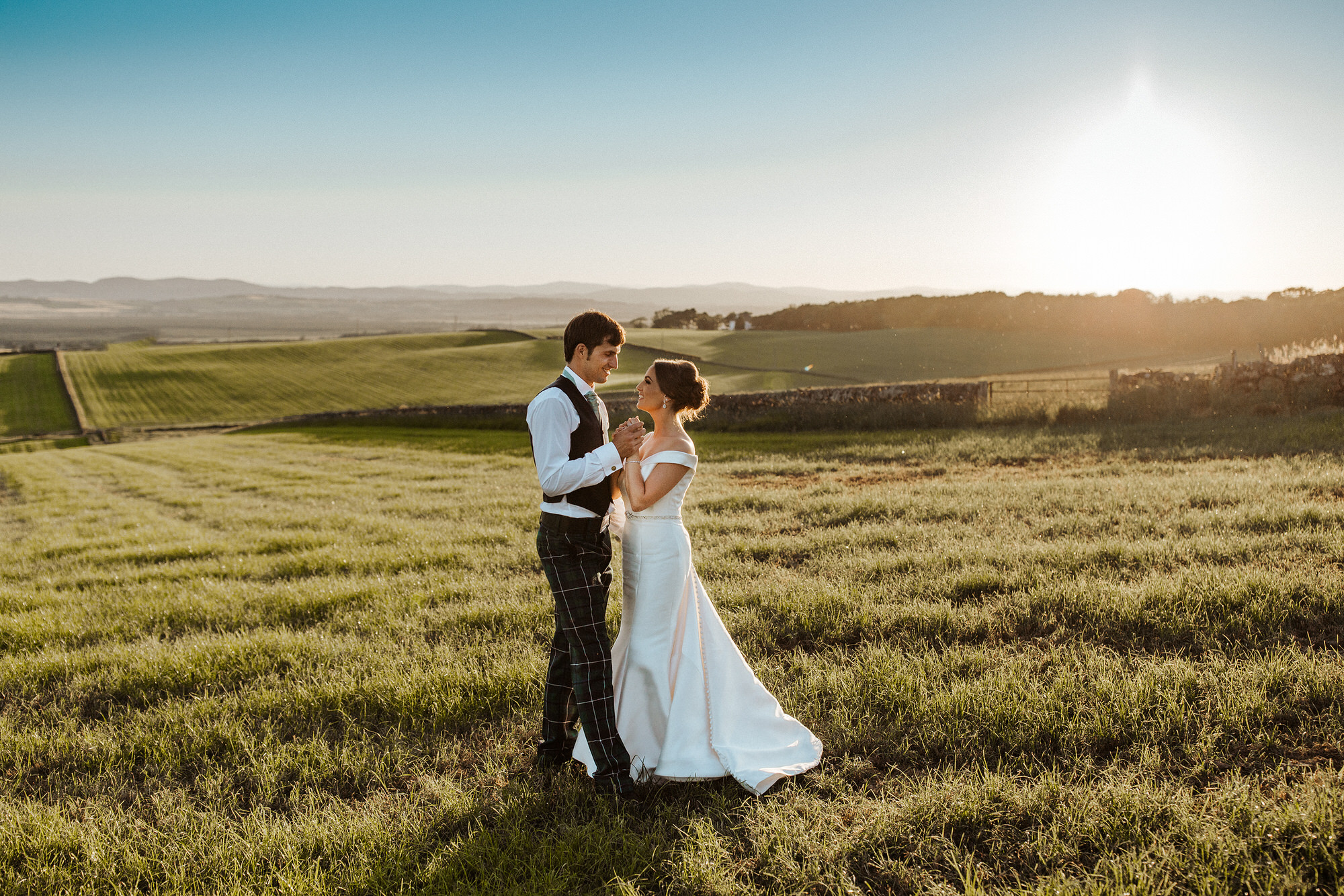 La mariée et le marié debout dans un champ vêtus d'une simple robe de mariée à épaules dénudées appelée Josie par Rebecca Ingram avec le marié dans un champ