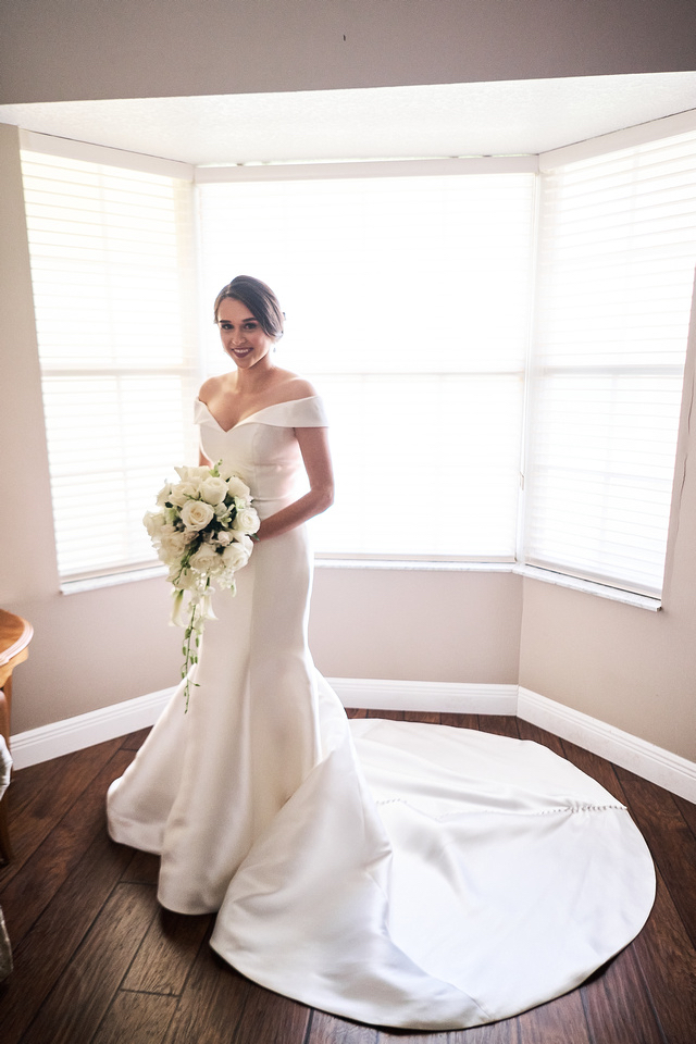 Bride Wearing Simple Off-The-Shoulder Wedding Dress Called Josie By Rebecca Ingram In Front Of Window
