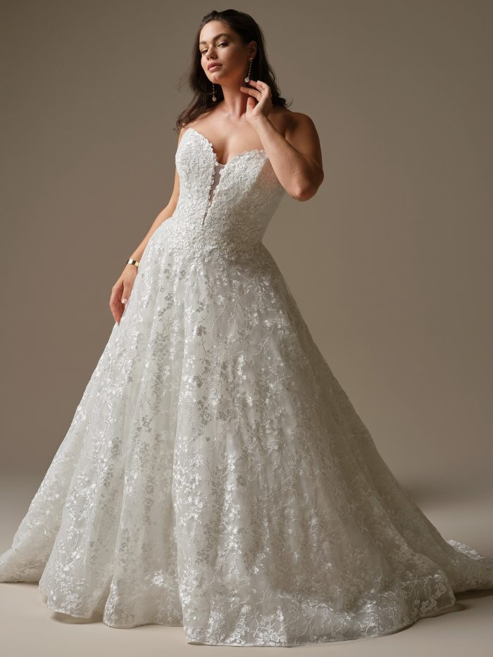Curve bride wearing the Memphis ballgown wedding dress by Maggie Sottero, featuring a strapless sweetheart neckline, plunging illusion detail, and shimmering floral lace over a full skirt.