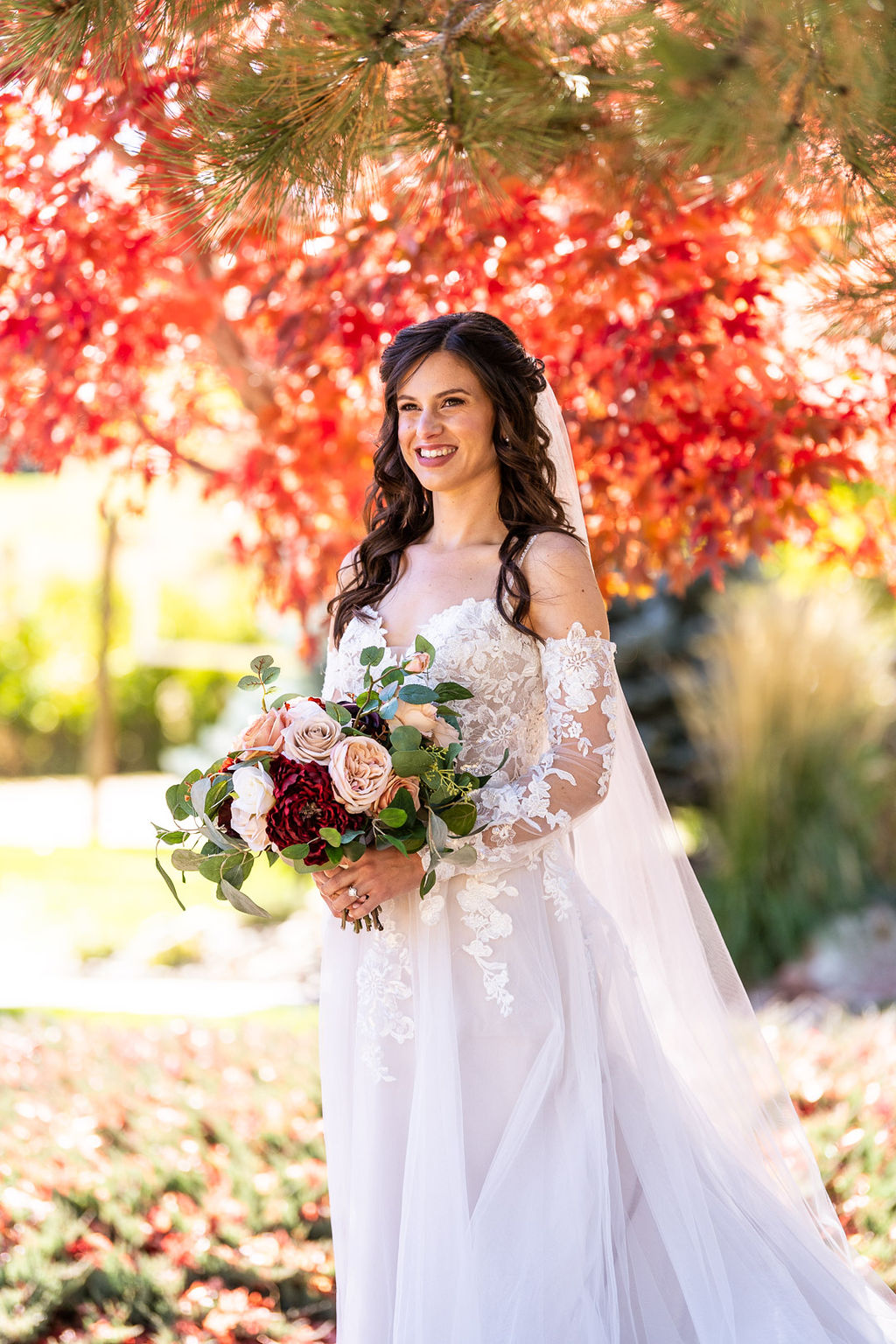Vraie mariée portant une robe de mariée à épaules dénudées devant des feuilles rouges à l'automne appelée Stevie par Maggie Sottero