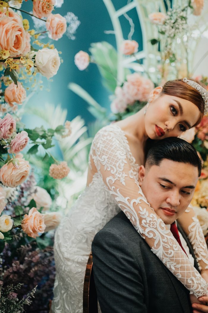 APPI Bride Wearing A Long Sleeve Wedding Gown Called Dakota With Chic Flower Wall Behind Her With Groom