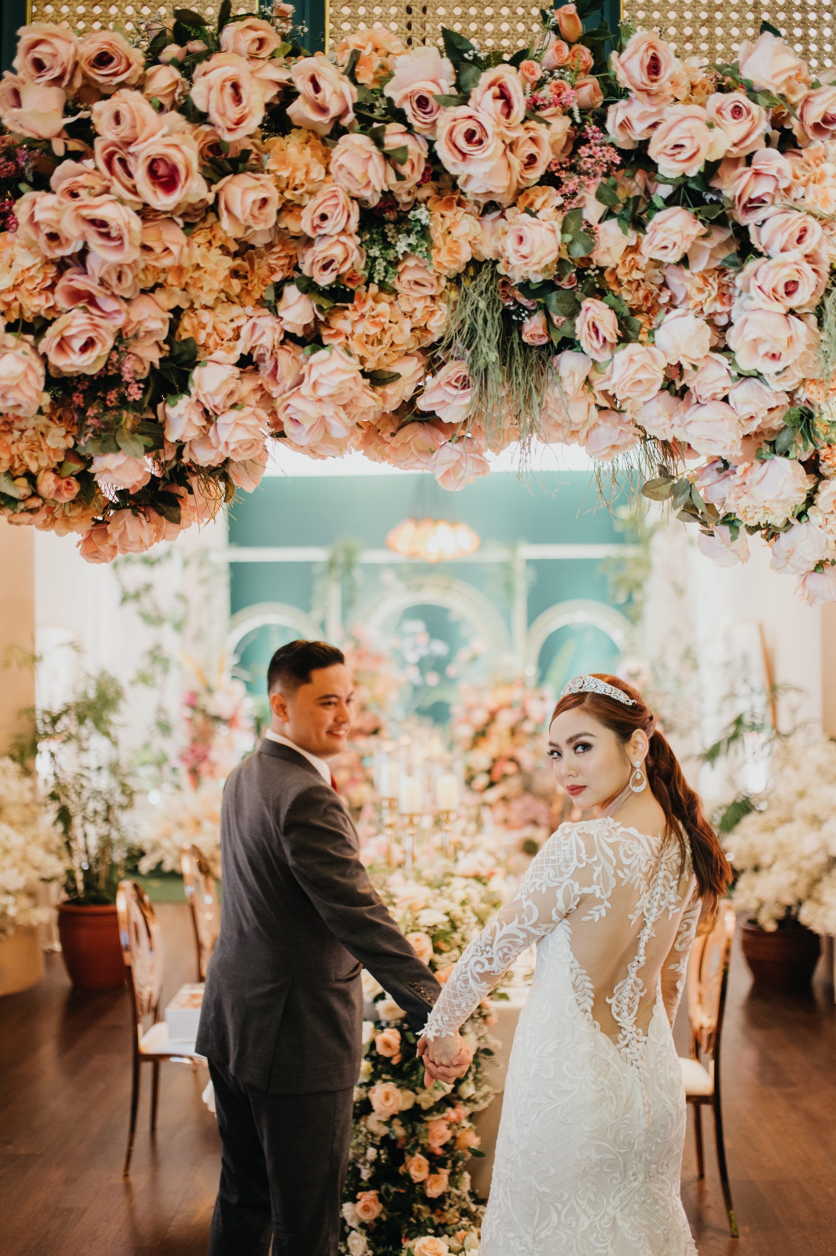 APPI mariée portant une robe de mariée à manches longues appelée Dakota avec un mur de fleurs chic derrière elle avec le marié