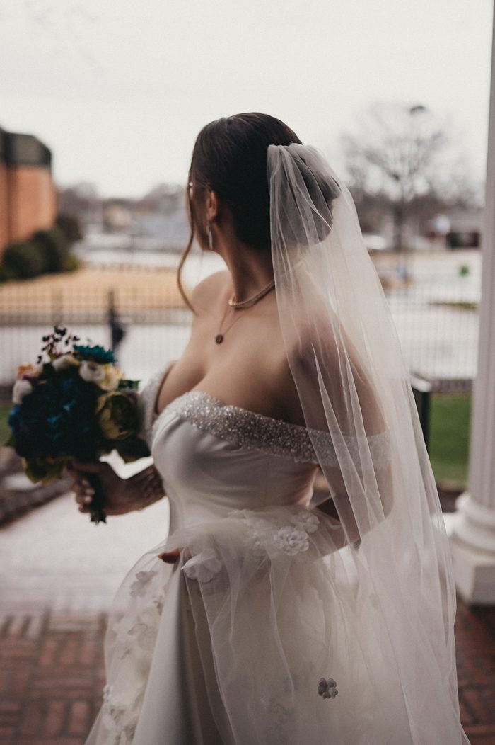 Bride Facing Away Holding Bridal Bouquet with a Veil and a Bun Hairstyle Wearing Fabienne Bridal Gown by Sottero and Midgley