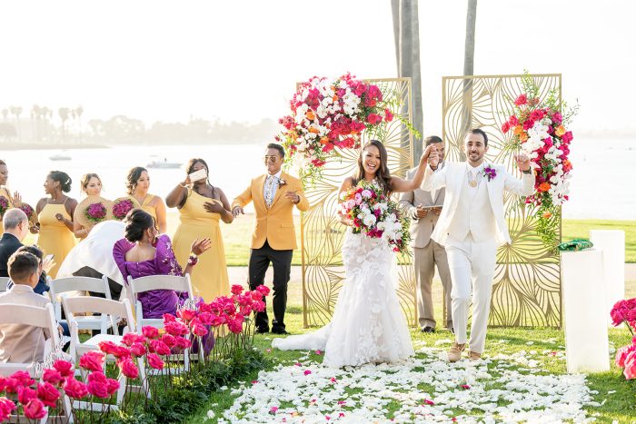 Bride In Floral Mermaid Wedding Dress Called Hattie By Rebecca Ingram With Groom In White Suit With Magenta Floral Wedding Arch