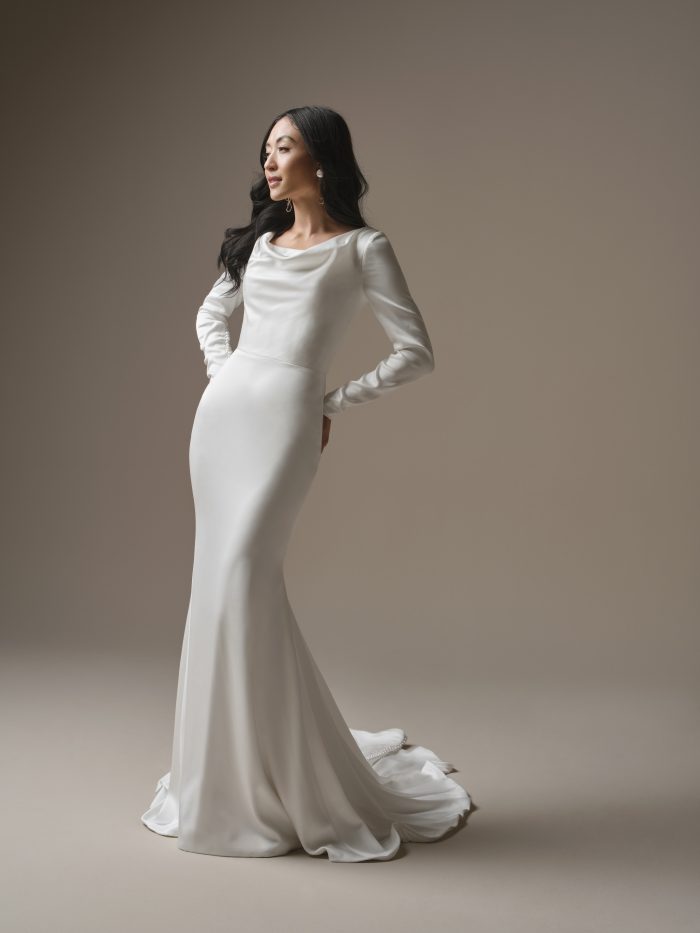 Bride wearing an ivory sheath wedding dress with a draped cowl neckline, long sleeves, fitted silhouette, and soft train.