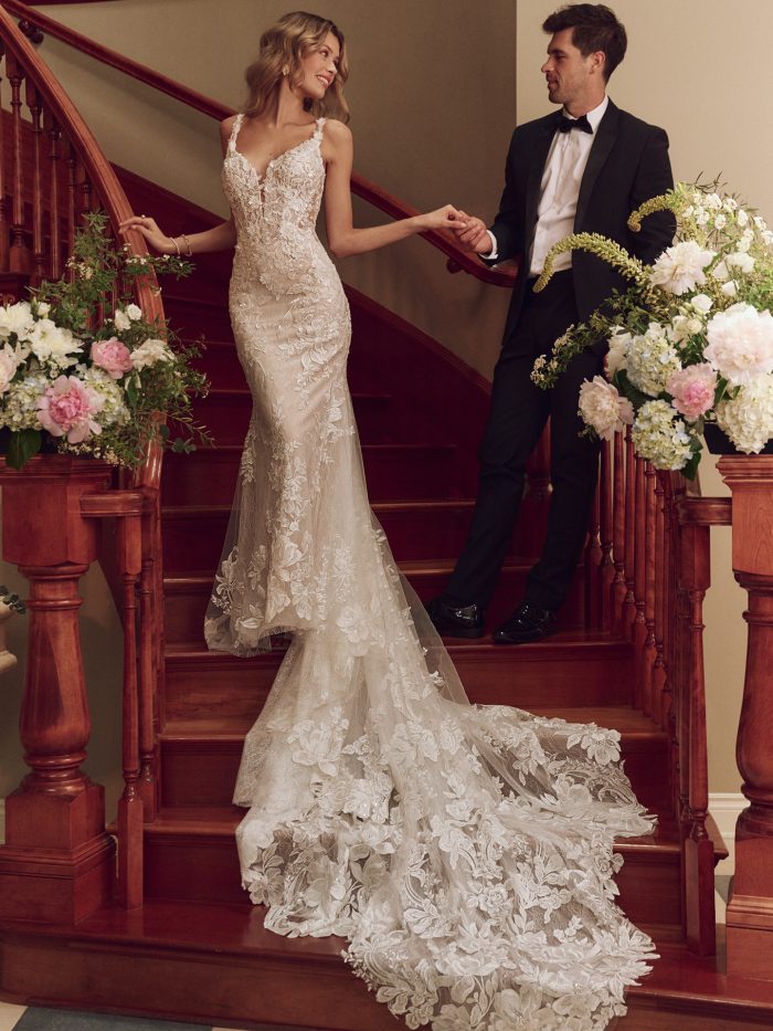 Bride in lace fit-and-flare wedding dress with illusion plunging neckline, floral appliqués, and dramatic cathedral train, styled on a grand staircase with groom.