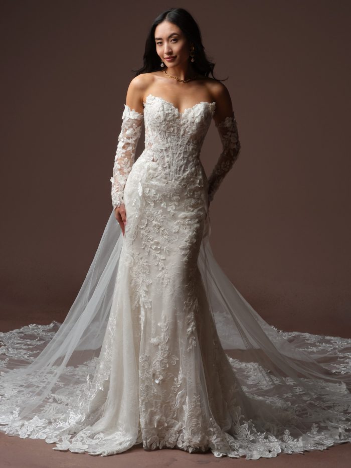 Bride wearing the Estelle fit-and-flare wedding dress, featuring an off-the-shoulder lace sweetheart neckline, sheer illusion long sleeves with floral lace appliqué, a fitted lace bodice and skirt, and a sheer overlay train with a scalloped lace edge.