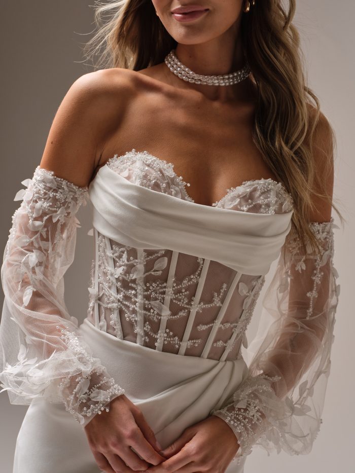 Bride wearing a strapless corset-style sheath wedding dress with an illusion beaded lace bodice, exposed boning, and sheer off-the-shoulder sleeves, paired with a draped crepe skirt.