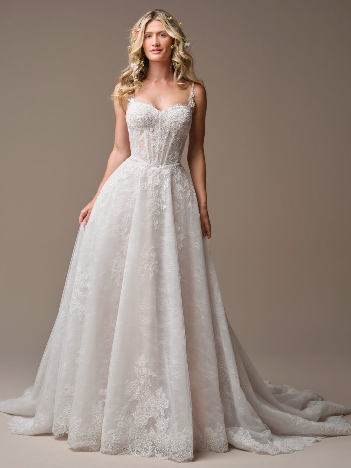 Bride wearing the Esmeralda A-line wedding dress, featuring a sweetheart neckline, corset-style bodice with sheer boning detail, thin lace straps, and a full lace-appliquéd tulle skirt with a scalloped hem and chapel train.