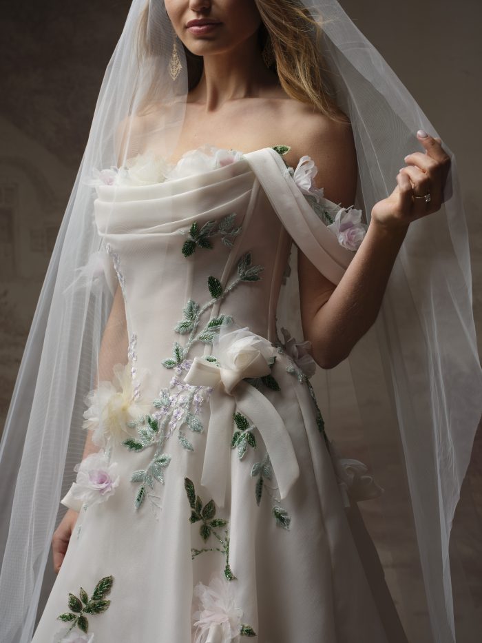 Close-up of the Amaryllis A-line wedding dress in blush, featuring a draped strapless neckline with 3D floral appliqués, beaded vine and leaf embroidery down the bodice, and a satin bow at the waist, worn with a sheer tulle veil.