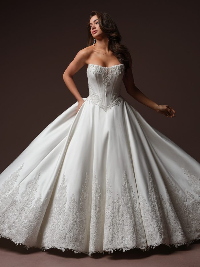 Bride wearing the Sandrine ballgown wedding dress, featuring a strapless sweetheart neckline with a boned corset bodice trimmed in beaded lace, and a dramatic full satin ballgown skirt with scalloped lace appliqué along the hem.