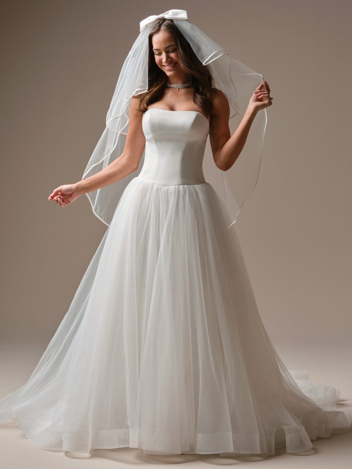 Model wearing the Shirley ball gown wedding dress, featuring a clean strapless satin bodice paired with a soft tulle skirt and styled with a bow-trimmed veil for a sweet, classic bridal look.