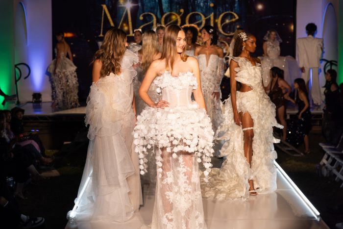 Runway show at Otis College of Art and Design featuring multiple models walking down a lit catwalk in layered, textured white gowns, with a “Maggie” backdrop and audience seated along the sides.