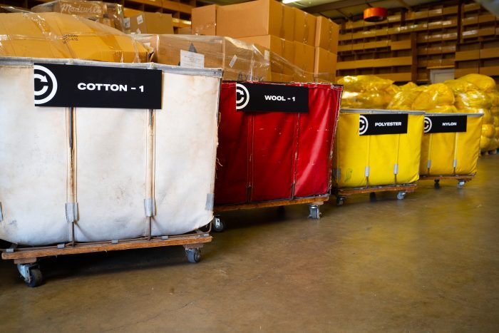 Large rolling bins in a warehouse labeled “Cotton - 1,” “Wool - 1,” “Polyester,” and “Nylon,” with stacked boxes and bundled materials in the background, representing textile sorting and recycling by SuperCircle.